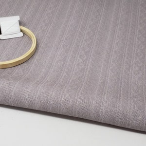May include: A close-up of a light purple fabric with a subtle vertical striped pattern. A wooden embroidery hoop and two spools of thread are on top of the fabric.