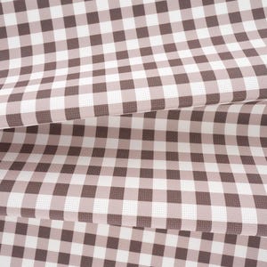 May include: A folded piece of fabric featuring a classic gingham pattern with alternating squares of dark brown and white. The textile has a visible woven texture and is draped in multiple layers, creating soft folds that highlight the grid design.