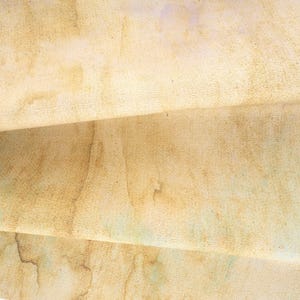May include: A piece of fabric featuring a textured, aged appearance with a light tan and beige base color. The surface displays irregular, mottled brown staining patterns and subtle hints of pale green and lavender hues across the weave.