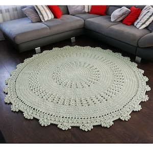 May include: A light green crocheted round rug with a delicate lace pattern. The rug is placed on a dark wood floor in front of a gray sectional sofa.