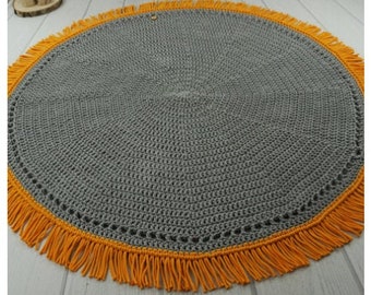 May include: A round gray crocheted rug with a yellow fringe border. The rug is made with a simple stitch pattern and has a subtle texture.