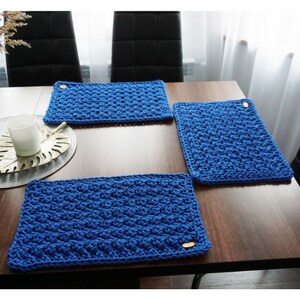 May include: Three blue crocheted placemats with wooden buttons on a wooden table.