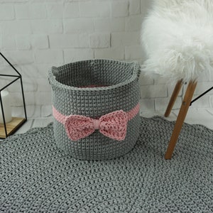 May include: A gray crocheted storage basket with a pink bow. The basket is sitting on a gray crocheted rug.