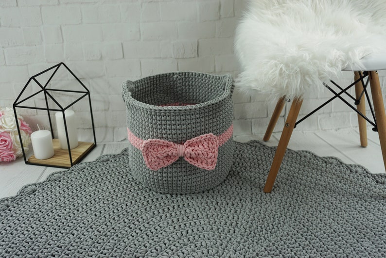 May include: A gray crocheted storage basket with a pink bow. The basket is sitting on a gray crocheted rug.