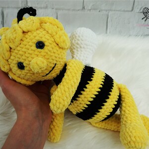 May include: A crocheted plush bee toy with yellow and black stripes, white wings, and black eyes. The bee is lying on its side with its head resting on a hand.