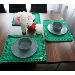 May include: Three green knitted placemats with a gold button detail on a wooden table. Each placemat has a white ceramic bowl and a gray ceramic plate on top.