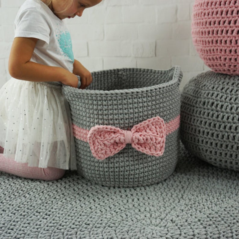 May include: A gray crocheted storage basket with a pink crocheted bow. The basket is sitting on a gray rug.