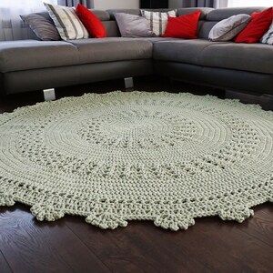 May include: A large, round, crocheted rug in a light green colour. The rug has a delicate, lacy pattern and is placed in front of a grey sectional sofa.