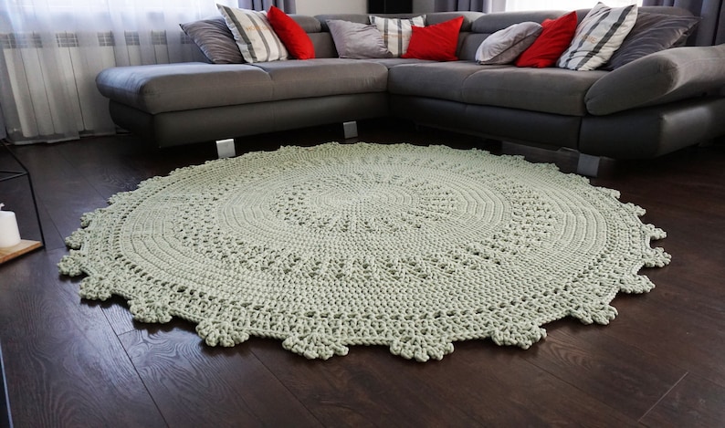 May include: A large, round, crocheted rug in a light green colour. The rug has a delicate, lacy pattern and is placed in front of a grey sectional sofa.