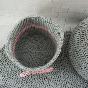 May include: A gray crocheted storage basket with a pink trim and a pink bow. The basket is sitting on a gray crocheted rug.