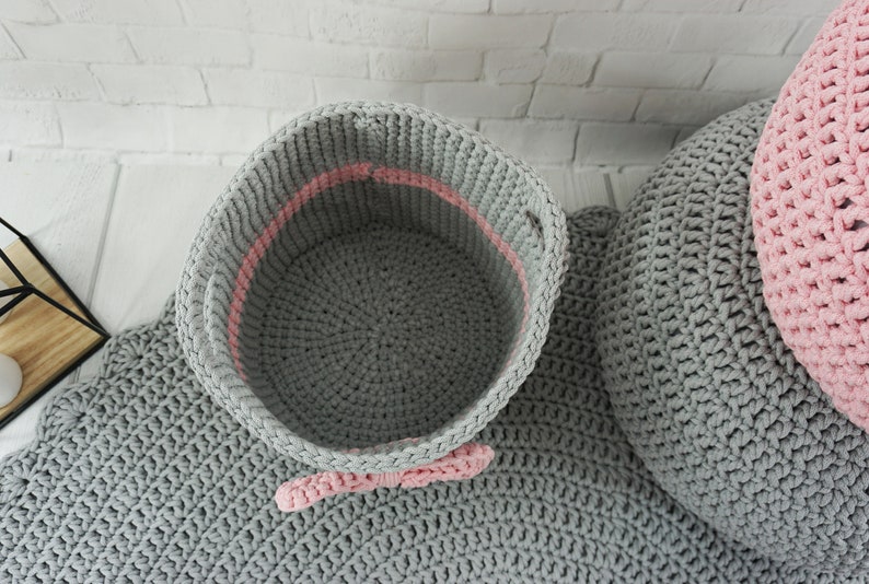May include: A gray crocheted storage basket with a pink trim and a pink bow. The basket is sitting on a gray crocheted rug.