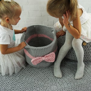 May include: Two children are playing with gray and pink crocheted storage baskets. The baskets have a pink ribbon bow on the front. The children are sitting on a gray crocheted rug.