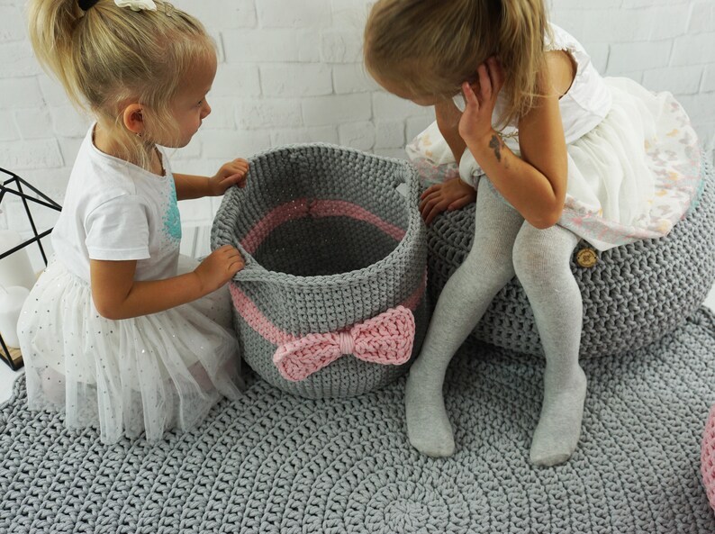 May include: Two children are playing with gray and pink crocheted storage baskets. The baskets have a pink ribbon bow on the front. The children are sitting on a gray crocheted rug.