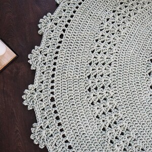 May include: A close-up of a light grey crocheted rug with a circular design. The rug has a delicate, lacy border.