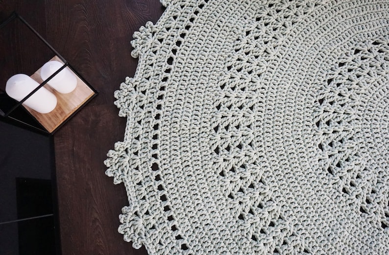 May include: A close-up of a light grey crocheted rug with a circular design. The rug has a delicate, lacy border.