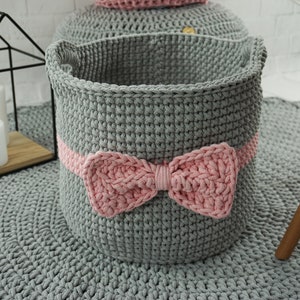 May include: A gray crocheted storage basket with a pink crocheted bow. The basket is sitting on a gray crocheted rug.