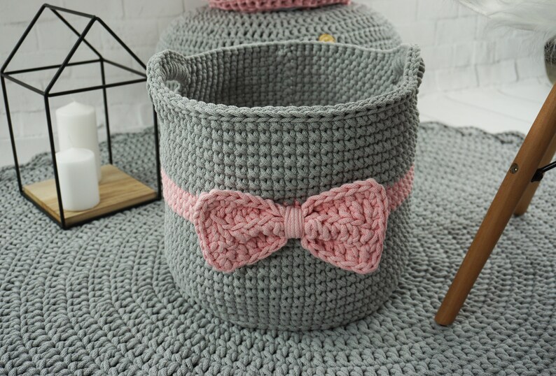 May include: A gray crocheted storage basket with a pink crocheted bow. The basket is sitting on a gray crocheted rug.
