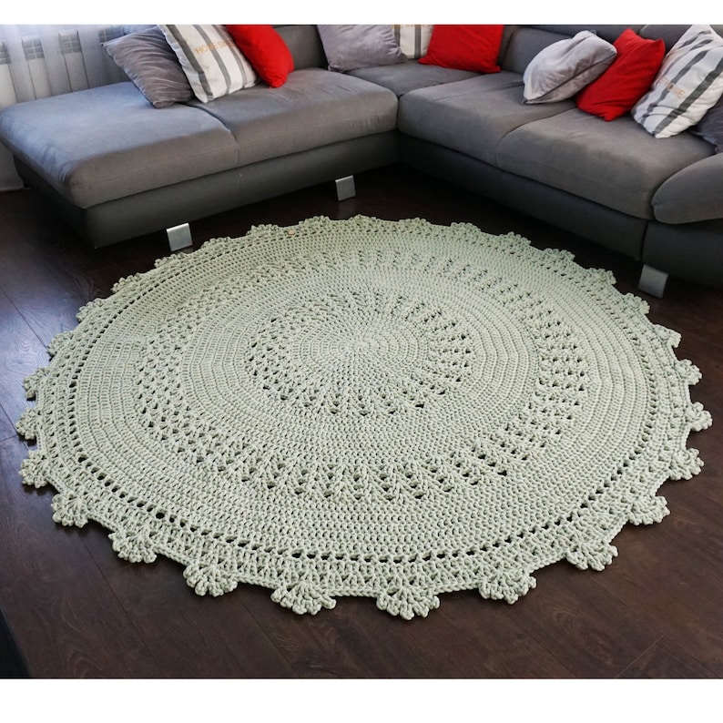 May include: A light green crocheted round rug with a delicate lace pattern. The rug is placed on a dark wood floor in front of a grey sectional sofa.