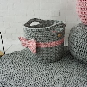 May include: A gray crocheted storage basket with a pink ribbon and a wooden button. The basket is sitting on a gray crocheted rug.