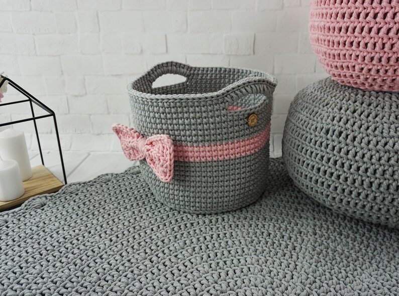 May include: A gray crocheted storage basket with a pink ribbon and a wooden button. The basket is sitting on a gray crocheted rug.