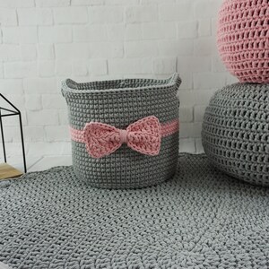 May include: A gray crocheted storage basket with a pink bow, sitting on a gray crocheted rug. There are two gray crocheted poufs in the background.
