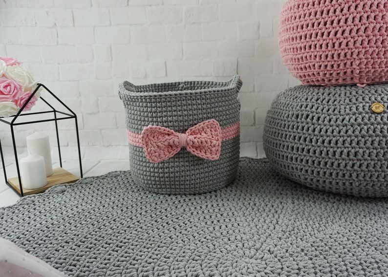 May include: A gray crocheted storage basket with a pink bow, sitting on a gray crocheted rug. There are two gray crocheted poufs in the background.