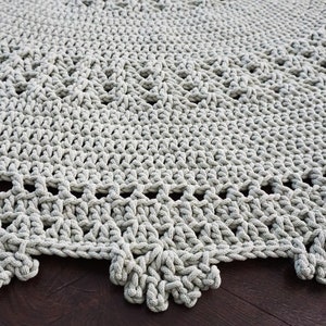 May include: A close-up of a grey crocheted rug with a scalloped edge. The rug is made with thick yarn and has a delicate pattern.