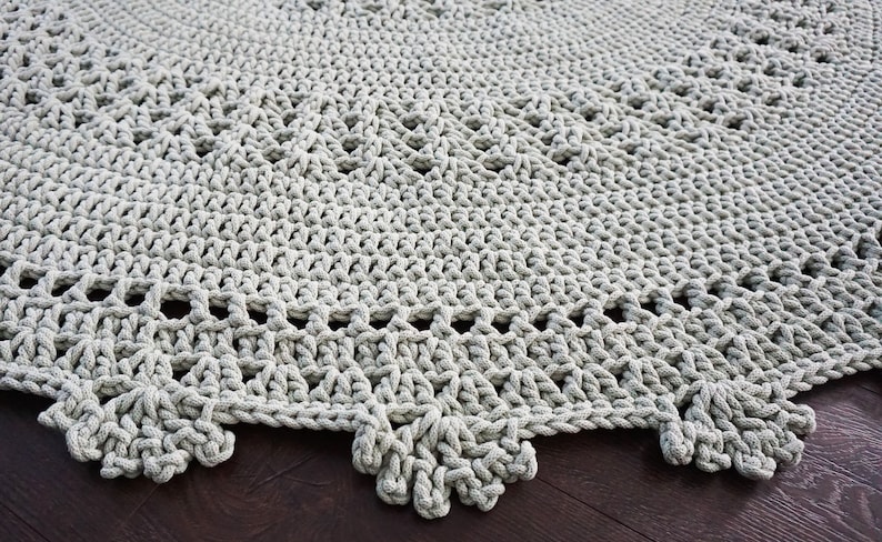May include: A close-up of a grey crocheted rug with a scalloped edge. The rug is made with thick yarn and has a delicate pattern.