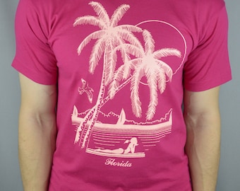 May include: A magenta short-sleeved t-shirt featuring a light pink graphic print of two palm trees, a sun, a bird, a sailboat, and a person lying on a surfboard. The word Florida is printed in a cursive script below the graphic illustration.