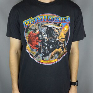 Pode incluir: Uma t-shirt preta de manga curta com uma impressão gráfica de um guerreiro a cavalo com uma capa vermelha e capacete. O texto MOLLY HATCHET aparece em azul e amarelo no topo, com o texto Lightning Strikes Twice na parte inferior.