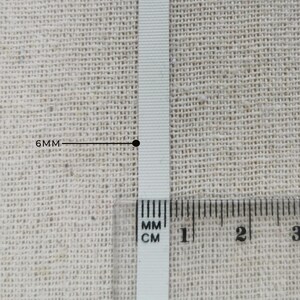 May include: A white ribbon with a ribbed texture positioned vertically. A clear plastic ruler is placed over the bottom section of the ribbon, and a black line with the text 6MM points to the width of the ribbon, indicating a 6mm measurement.