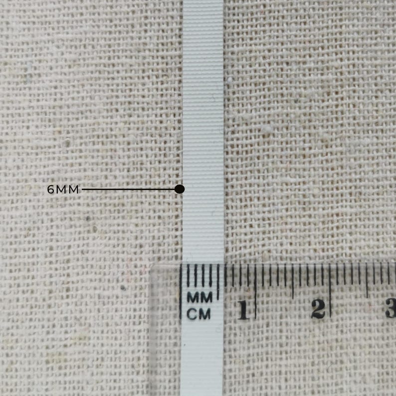 May include: A white ribbon with a ribbed texture positioned vertically. A clear plastic ruler is placed over the bottom section of the ribbon, and a black line with the text 6MM points to the width of the ribbon, indicating a 6mm measurement.
