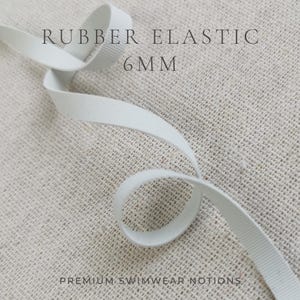 May include: A white rubber elastic band measuring 6mm in width, featuring a ribbed texture throughout its length. The elastic is coiled in a loose loop. The text RUBBER ELASTIC 6MM and PREMIUM SWIMWEAR NOTIONS is printed on the image.