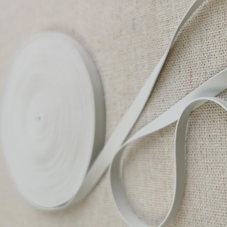 May include: A spool of light gray ribbon featuring a fine, ribbed texture. The ribbon is wound into a tight, circular coil, with a loose end extending outward, showcasing the consistent width and the subtle, woven pattern of the material.