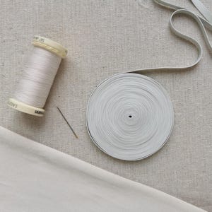 May include: A spool of light beige sewing thread, a single silver sewing needle, and a circular roll of flat white elastic tape. The items are arranged on a textured, light-colored fabric surface, showcasing essential sewing supplies.