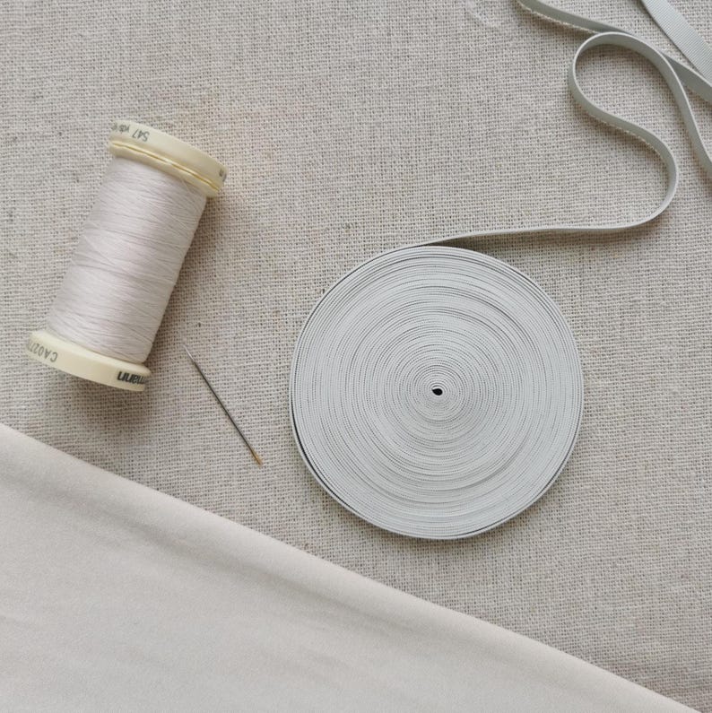 May include: A spool of light beige sewing thread, a single silver sewing needle, and a circular roll of flat white elastic tape. The items are arranged on a textured, light-colored fabric surface, showcasing essential sewing supplies.