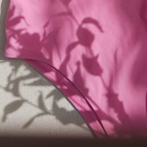May include: A solid bright pink fabric garment featuring a curved hemline with visible double-stitched seams. The surface of the pink material is partially obscured by the dark, intricate shadows of leaves and flowers cast across the fabric.