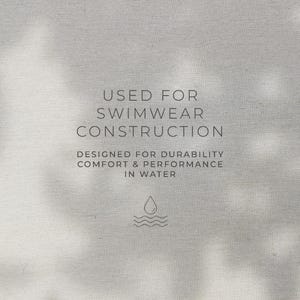 May include: A light gray fabric background featuring dark gray text that reads: USED FOR SWIMWEAR CONSTRUCTION. DESIGNED FOR DURABILITY COMFORT & PERFORMANCE IN WATER. Below the text is a small icon of a single water droplet above three wavy lines.