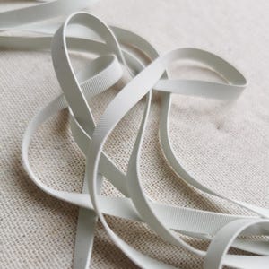 May include: A length of white grosgrain ribbon featuring a distinct horizontal ribbed texture. The narrow, flexible fabric strip is coiled in loose loops, showcasing its consistent matte finish and uniform width throughout the entire length.