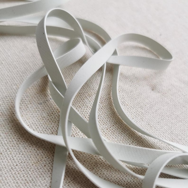May include: A length of white grosgrain ribbon featuring a distinct horizontal ribbed texture. The narrow, flexible fabric strip is coiled in loose loops, showcasing its consistent matte finish and uniform width throughout the entire length.