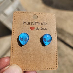 May include: A pair of blue alien-shaped stud earrings. The earrings are on a brown card that reads "Handmade with love." The alien heads have black eyes and a shiny, iridescent finish.