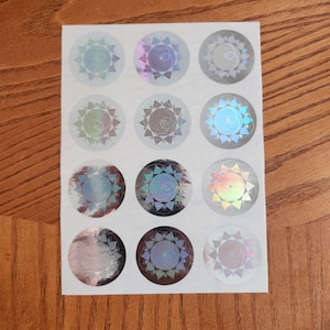 Op de afbeelding: Een vel met 12 regenboogkleurige holografische stickers met een geometrisch stervormig ontwerp. Elke sticker heeft een andere kleurencombinatie van blauw, groen, paars en oranje.