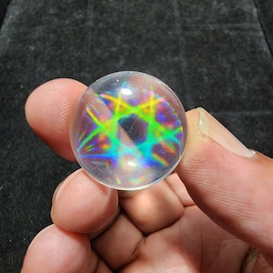 Puede incluir: Una esfera de vidrio transparente con colores arcoíris iridiscentes en su interior. La esfera es del tamaño de una canica y se está sosteniendo en una mano.