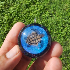 May include: A round pendant featuring a detailed sea turtle design. The turtle is brown and tan, set against a blue, glittery background. The pendant has a black border with colorful glitter accents and a silver loop for a chain.