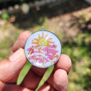 May include: A round pendant with a colorful design featuring a sun, a figure, mushrooms, and swirling pink clouds. The pendant has a silver-colored bail for attaching to a chain. The design has a retro, psychedelic vibe.