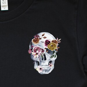 May include: Black t-shirt with a white skull design. The skull's eye sockets are filled with watercolor flowers in shades of pink, purple, yellow, and brown.