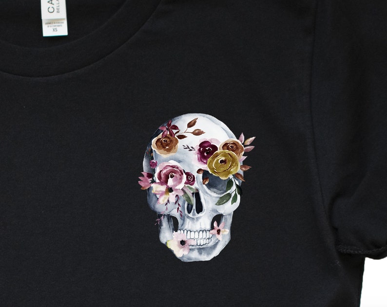 May include: Black t-shirt with a white skull design. The skull's eye sockets are filled with watercolor flowers in shades of pink, purple, yellow, and brown.