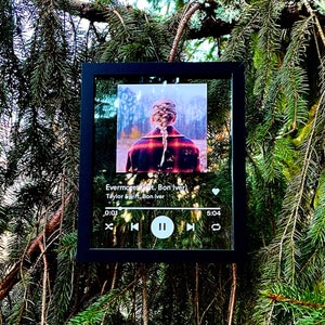 Op de afbeelding: Een ingelijste glazen print van een muziekspeelerscherm waarop het nummer "Evermore" van Taylor Swift met Bon Iver te zien is. De afbeelding toont een vrouw met gevlochten haar die een rood-zwart geruit overhemd draagt.