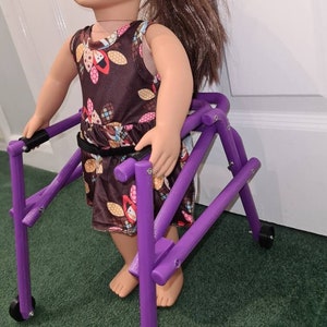 Puede incluir: Un andador de muñeca morado con ruedas. El andador está hecho de plástico y tiene un diseño simple. Una muñeca está de pie junto al andador, sujetando las asas.