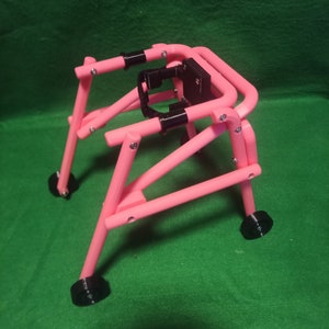 Puede incluir: Un andador de plástico rosa con ruedas negras y un marco de plástico negro. El andador está diseñado para ayudar a los niños a aprender a caminar.